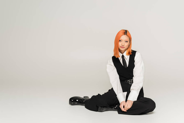 full length of confident and successful asian woman sitting and looking at camera on grey background, business casual, black vest, tie and pants, white shirt, colored red hair, youthful style - Photo, Image