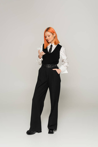 full length of trendy asian woman with dyed red hair standing with hand in pocket and networking on mobile phone on grey background, business casual fashion, white shirt, black vest and pants - Foto, Bild