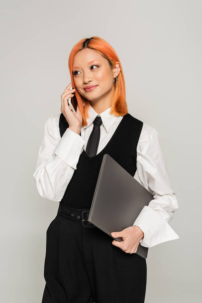 happy emotion, phone call, young asian woman holding laptop and smiling during mobile communication on grey background, colored red hair, white shirt, black vest and tie, business casual fashion - Fotó, kép