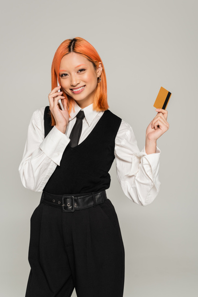 online shopping, excitement, red haired asian woman with credit card making order on smartphone on grey background, business casual style, white shirt, black vest and tie, generation z - Photo, Image