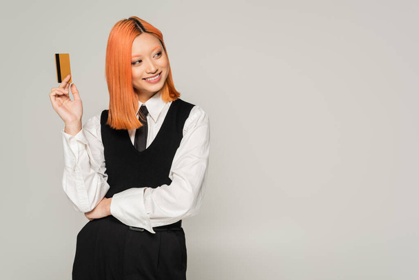happy emotion, cheerful smile, red haired asian woman holding credit card and looking away on grey background, business casual style, white shirt, black tie and vest, generation z - Photo, Image