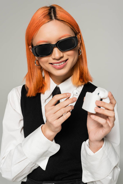 сверкающая азиатская женщина с сияющей улыбкой и цветными рыжими волосами, держащая футляр с беспроводными наушниками на сером фоне, деловой повседневный стиль, темные солнечные очки, белая рубашка, черный галстук и жилет - Фото, изображение