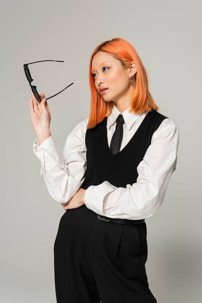 business casual fashion, attractive asian woman with colored red hair, in white shirt, black pants, vest and tie holding dark sunglasses and looking away on grey background, generation z - Photo, Image