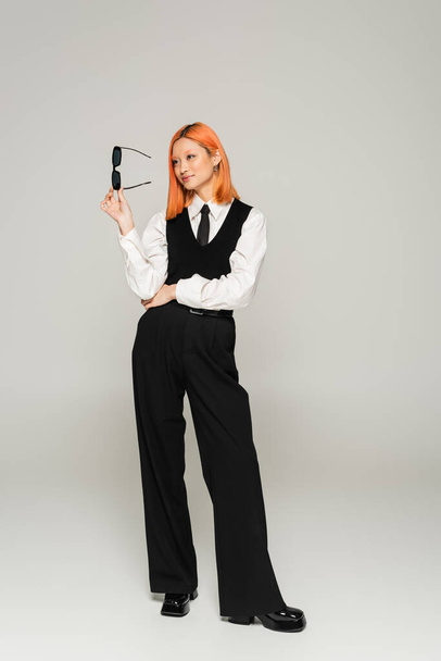 happy emotion, full length of asian woman with dyed red hair holding dark sunglasses and looking away on grey background, business casual fashion, white shirt, black tie, vest and pants - Valokuva, kuva