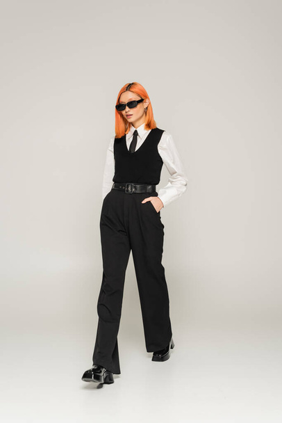 full length of stylish asian woman with dyed red hair, in dark sunglasses walking with hand in pocket on grey background, business fashion, white shirt, vest and pants, modern lifestyle - Foto, imagen