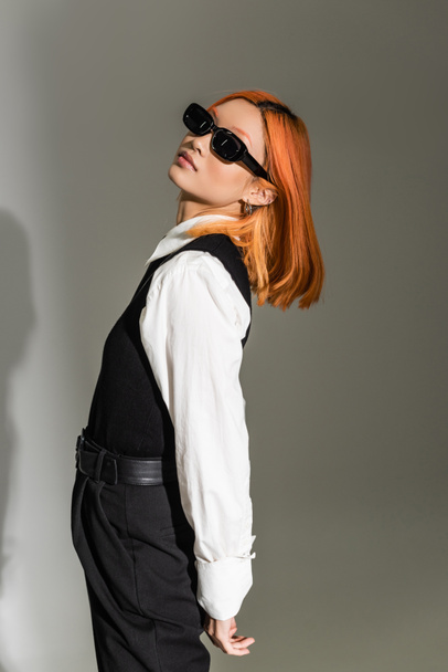 charming and red haired asian woman in dark stylish sunglasses, white shirt and black vest standing and looking at camera on grey shaded background, business casual, generation z lifestyle - Photo, Image
