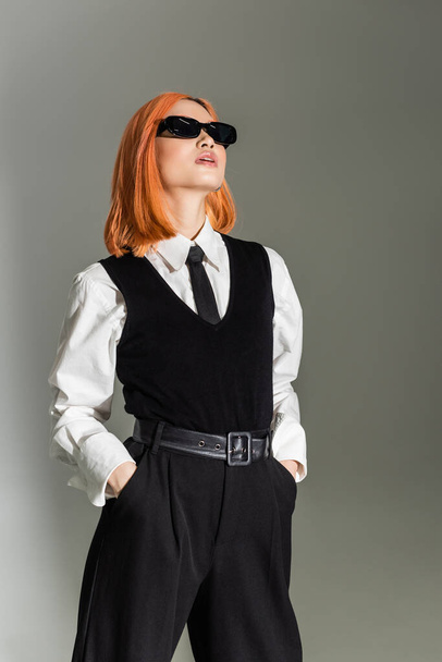 confident asian woman in dark sunglasses standing with hands in pockets and looking away on grey shaded background, business casual, white shirt, black tie, vest and pants, modern fashion - Foto, Imagem