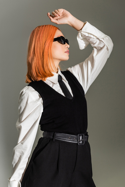 side view of expressive and stylish asian woman posing with hand above head on grey shaded background, dyed red hair, dark sunglasses, white shirt, tie and vest, business casual fashion - Zdjęcie, obraz