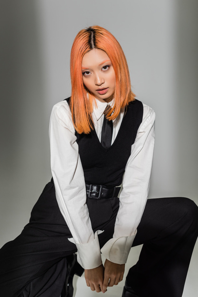 young and expressive asian woman in black and white business casual clothes looking at camera on grey shaded background, colored red hair, white shirt, black tie and pants, modern fashion - Fotó, kép