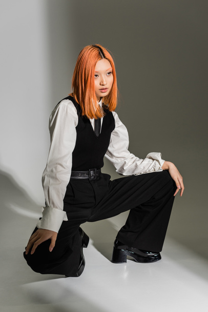 red haired and confident asian woman in stylish business clothes posing and looking away on grey shaded background, white shirt, black vest and pants, modern fashion, generation z - Photo, Image