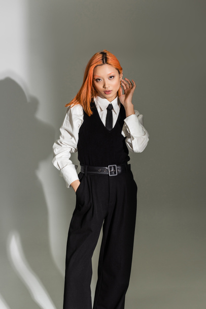 trendy and confident asian woman touching red colored hair, holding hand in pocket and looking at camera on grey shaded background, business casual, white shirt, black tie, shirt and pants - Foto, afbeelding