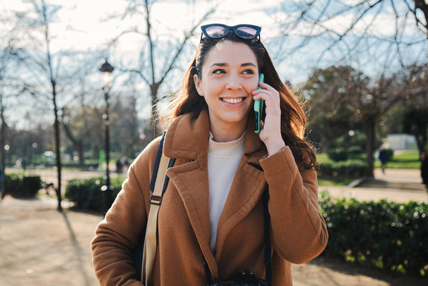 Happy young woman smiling having a friendly call conversation with a smartphone. Caucasian female talking by cellphone. Cheerful lady using a mobile phone device to communicate standing outdoors. High - Photo, Image