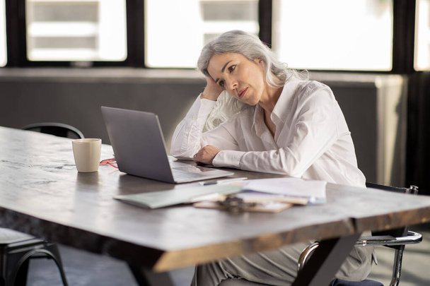 Professional Burnout. Tired Mature Businesswoman Looking At Laptop, Exhasuted After Stressful Online Work, Sitting At Desk In Modern Office Interior. Workplace Problems And Fatigue Concept - Fotoğraf, Görsel