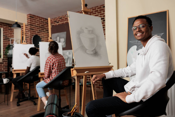 Portrait of happy young African American man student of art school sitting at easel smiling at camera, learning pencil drawing in class. Positive black guy enjoying creative hobby with friends - Photo, Image