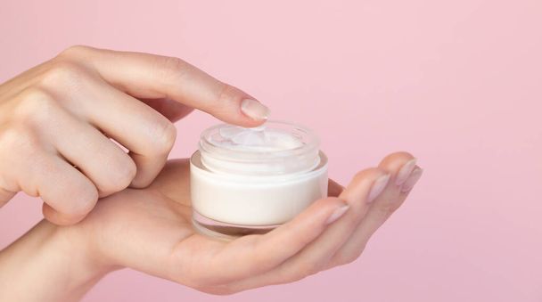Beautiful graceful fingers of a woman scoop cream from a round glass jar on a pink background. Hand care concept. Layout for cosmetic products - Photo, Image