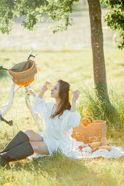 Hermosa joven morena en un picnic en el campo. Modelo en ropa elegante con bicicleta. - Foto, imagen