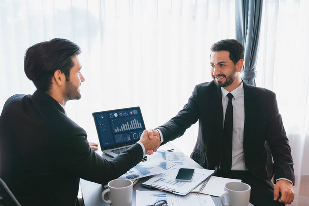 Closeup professional businessman shaking hands over desk in modern office after successfully analyzing pile of dashboard data paper as teamwork and integrity handshake in workplace concept. fervent - Photo, Image