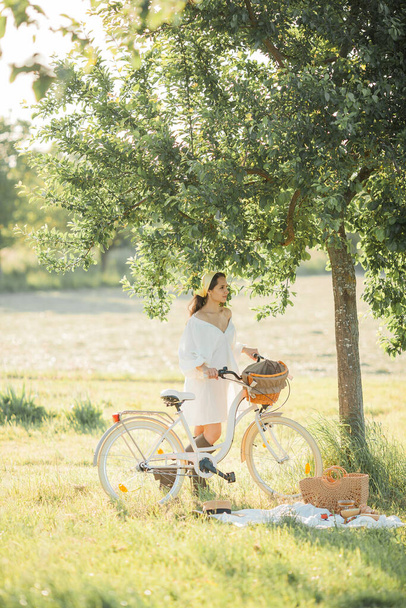 Όμορφη νεαρή μελαχρινή γυναίκα σε πικ-νικ στο πεδίο. Μοντέλο σε κομψά ρούχα με ποδήλατο. - Φωτογραφία, εικόνα