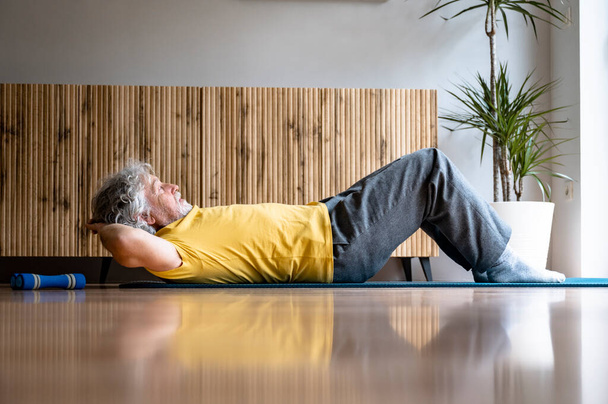 Uomo anziano in camicia gialla che lavora fuori praticando un esercizio di pilates per i muscoli del nucleo forte un esercizio di sit up crunch su un tappeto. - Foto, immagini