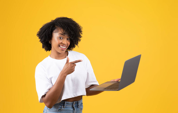Happy millennial african american curly woman in white t-shirt pointing finger at laptop, isolated on yellow studio background. Recommendation device, social media, app and blog, work and study - Фото, зображення
