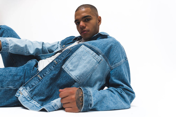 Homem afro-americano vestindo um casaco de ganga e jeans marcando uma pose no chão. Isolado contra um fundo branco. Imagens 4k de alta qualidade - Foto, Imagem