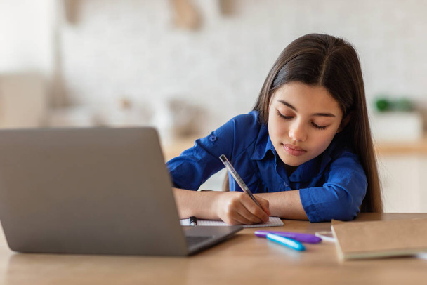 Διαδικτυακό σχολείο. Μαθήτρια που κρατάει σημειώσεις γράφοντας σε ένα βιβλίο κοντά στο Laptop και μελετώντας από το σπίτι, καθισμένη σε ένα γραφείο. Απομακρυσμένη Εκπαίδευση Ελεύθερος χρόνος, Σύγχρονη Εκπαίδευση και Τεχνολογία Διαδικτύου - Φωτογραφία, εικόνα