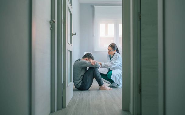 Γυναίκα γιατρός καθησυχάζει και βοηθά με την ενσυναίσθηση σε αρσενικό ασθενή κάθεται στο πάτωμα του δωματίου ενός κέντρου ψυχικής υγείας - Φωτογραφία, εικόνα