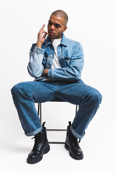 Black model in denim clothes posing on a black folding chair against a white background. High-quality 4k footage - Photo, Image