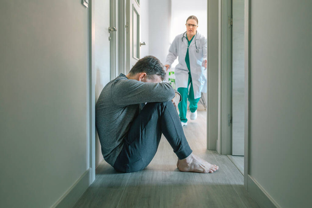Беспокойная женщина-врач бежит, чтобы помочь пациенту с психическими расстройствами и суицидальными мыслями, сидя на полу коридора - Фото, изображение