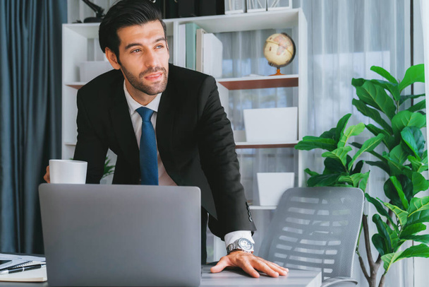 Современный профессиональный бизнесмен за современным офисным столом использует ноутбук для работы с кофе в руке. Трудолюбивый офисный работник, работающий над ноутбуком в своем офисе. пылкий - Фото, изображение