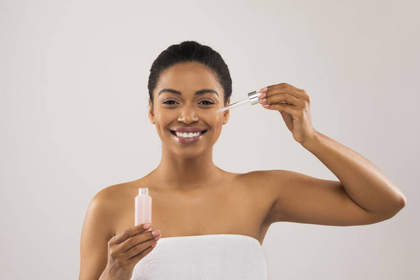 Smiling happy pretty young black woman wrapped in towel holding small bottle with beauty product, using anti-aging hydrating serum or nourishing oil for youthful looking skin, studio background - Photo, Image