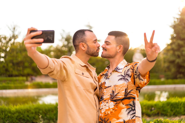 Homosexual couple taking a selfie and kissing at sunset in a park in the city. Diversity and lgbt concept - Foto, immagini