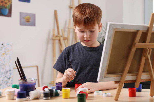 Little boy painting at table in studio. Using easel to hold canvas - Фото, изображение