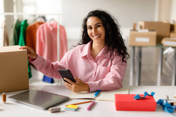 Επιχειρήσεις κατάστημα ρούχων. Ευτυχισμένο Μεσανατολίτικη Manager Lady Χρησιμοποιώντας Smartphone Και Συσκευασίας Παραγγελία Ρούχα Σε Χάρτινο Κουτί, Ιδιοκτησία Online Κατάστημα Μόδας, Κάθεται Στο Γραφείο Εσωτερικού Χώρου. Λιανικό και ηλεκτρονικό εμπόριο - Φωτογραφία, εικόνα
