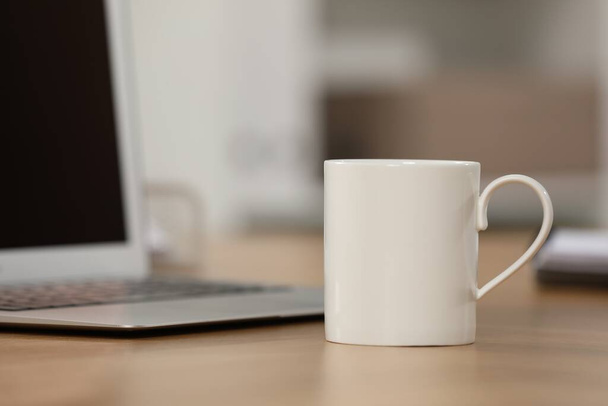 White ceramic mug and laptop on wooden table at workplace. Space for text - Foto, Imagem