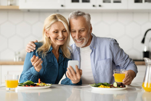 キッチンで朝食中にスマートフォンを使用して幸せなシニア配偶者の肖像,携帯電話でビデオ通話を行う夫婦や新しいアプリをチェック,無料のスペースで終了ショット - 写真・画像