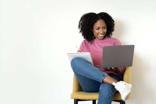 Busy happy millennial african american curly lady in casual sits on armchair, typing at computer and makes notes on white wall background. Work and study, video lesson, webinar, education at home - Fotoğraf, Görsel