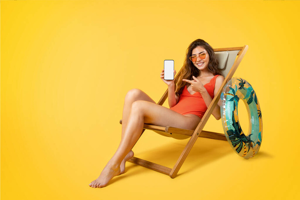 Jovem alegre senhora de raça mista em maiô, óculos de sol sentar na cadeira deck, dedo apontador no telefone com tela em branco, desfrutar de férias, isolado no fundo amarelo. App para férias no verão, blog - Foto, Imagem