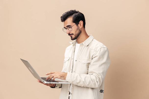 Портрет стильного задумчивого мужчины в очках с ноутбуком в руках фрилансера, на бежевом фоне в белой футболке, модный стиль одежды, пространство. Высокое качество фото - Фото, изображение