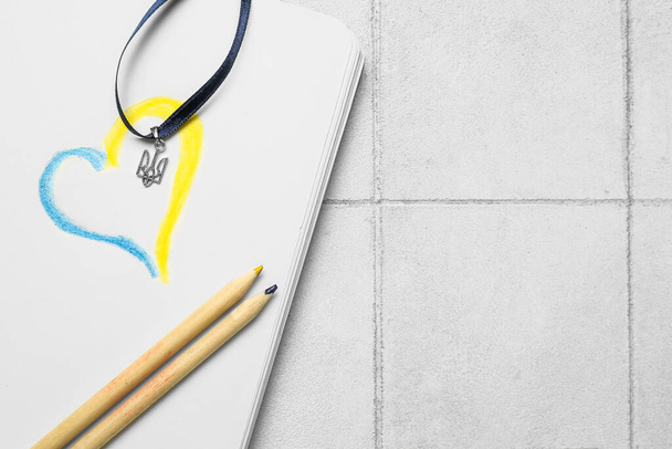 Ribbon with Ukrainian coat of arms, notebook, drawn heart and pencils on white tile background - Fotó, kép