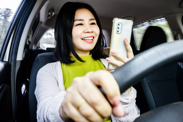 zorgeloze aziatische vrouw met behulp van een mobiele telefoon en niet het dragen van een gordel tijdens het rijden op de weg, Risky rijgewoonten concept - Foto, afbeelding