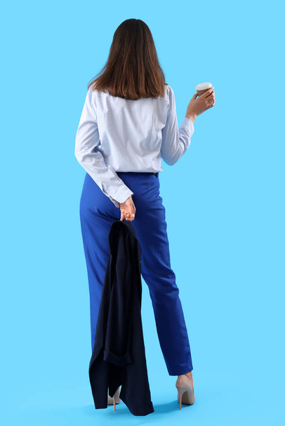 Mujer joven en traje elegante con taza de café sobre fondo azul, vista trasera - Foto, Imagen