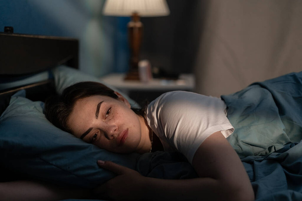 Jonge verdrietige vrouw die 's avonds laat in bed ligt en probeert te slapen terwijl ze slapeloosheid lijdt. Meisje in bed bang voor nachtmerries die er bezorgd en gestrest uitzien. Slaapstoornis en slapeloosheid concept - Foto, afbeelding