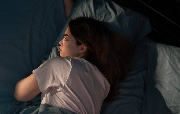 Молодая грустная женщина, лежащая в постели поздно ночью, пытаясь уснуть, страдая бессонницей, выглядит обеспокоенной. Концепция психического здоровья. Вид сверху - Фото, изображение