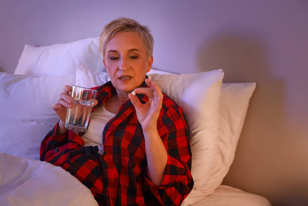 Mature woman with glass of water and pill in bed at night - Photo, Image