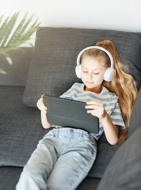 Kanepede elektronik tabletle uzanmış küçük bir kız beyaz kulaklıkla müzik dinliyor. Eğitim için kablosuz aygıtlar kullanılıyor. - Fotoğraf, Görsel