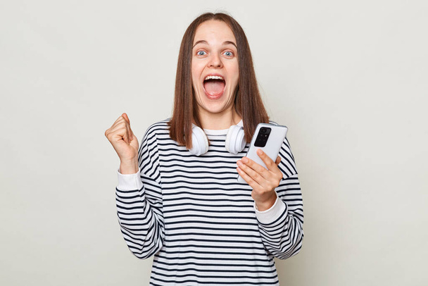 Extremely happy amazed brown haired woman wearing striped shirt posing isolated over gray background standing with clenched fist holding mobile phone gets sms. - Фото, изображение