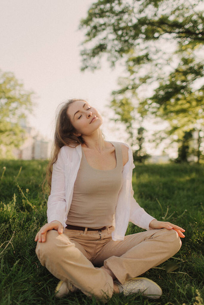 Йога в парке с солнечным светом. Молодая женщина в позе лотоса сидит на зеленой траве. Концепция спокойствия и медитации. Понятие здорового образа жизни. - Фото, изображение
