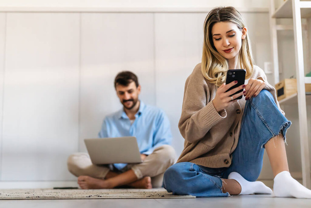 Усміхнена молода жінка використовує смартфон з чоловіком, який сидить з ноутбуком. Люди працюють в соціальних мережах концепція домашнього офісу
. - Фото, зображення
