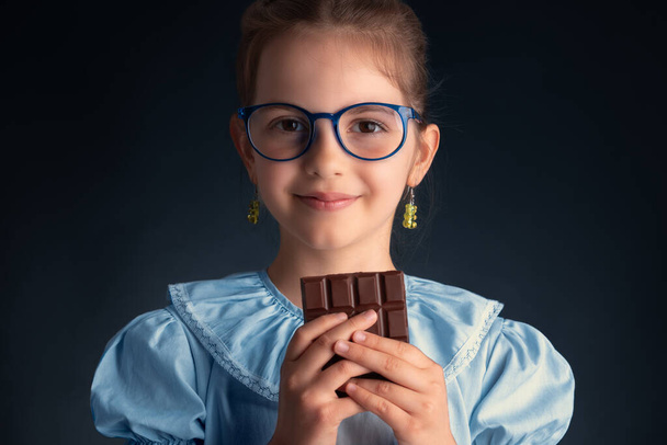 Chocolate and pretty hungry little woman portrait. Beautiful clever girl with glasses ready to eat chocolate bar. - Photo, Image